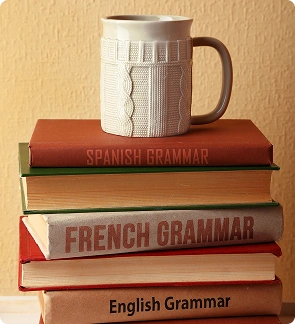 Een witte, gestructureerde mok staat bovenop een stapel grammatica boeken: Spaans, Frans en Engels. De scène straalt een gezellige, studieuze sfeer uit.