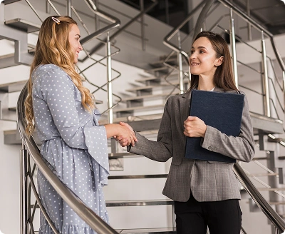 Twee professionele vrouwen die elkaar de hand schudden in een modern kantoor bij een trap, wat partnerschap, vertrouwen en succesvolle zakelijke samenwerking symboliseert.