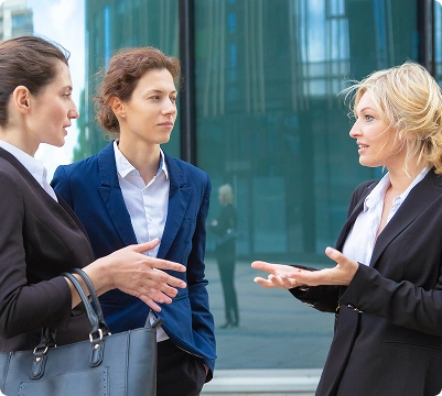 Drie professionele zakenvrouwen die buiten een modern kantoorgebouw een discussie hebben, wat communicatie, teamwork en strategische planning vertegenwoordigt.